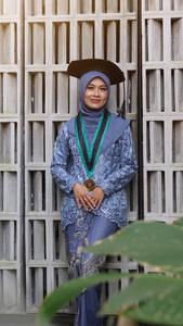 A smiling woman in graduation attire stands against a wall.